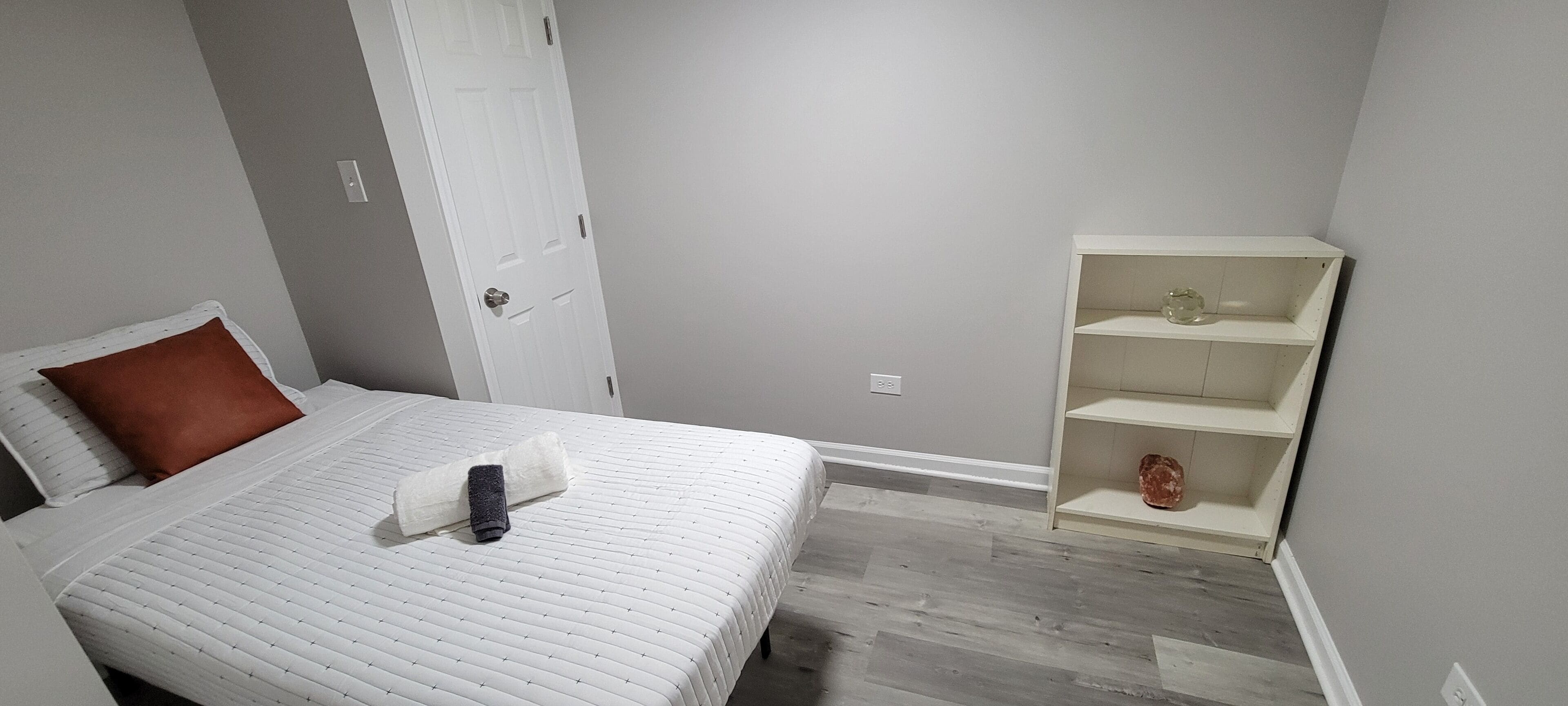 A bedroom with a bed, a white door, a white shelf unit, and a grey wall.