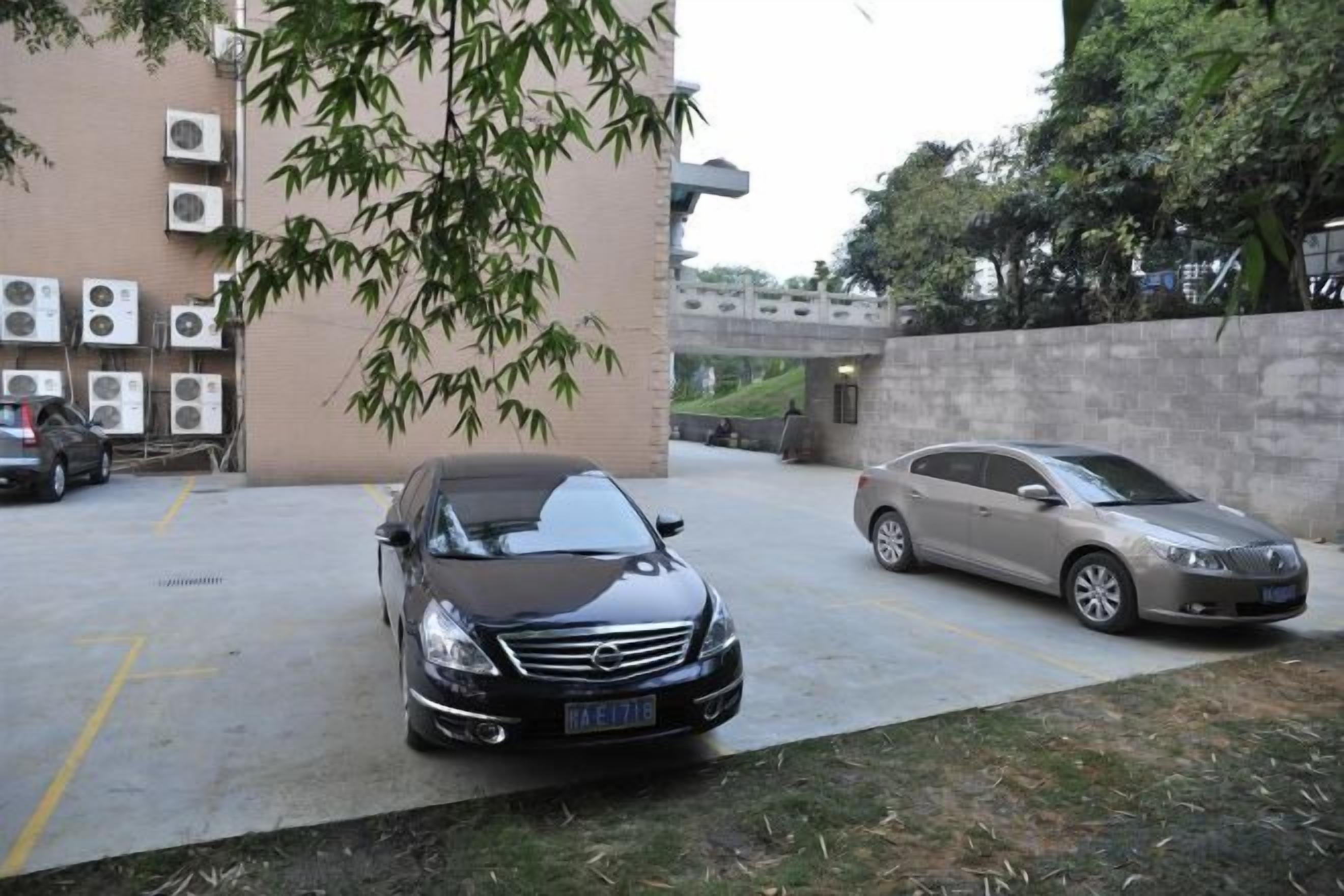 Un área de estacionamiento con dos coches, un edificio con unidades de aire acondicionado y un árbol.
