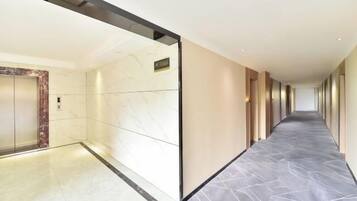 A long, narrow hallway with a marble floor, a wall-mounted elevator, and a door with a sign above it.