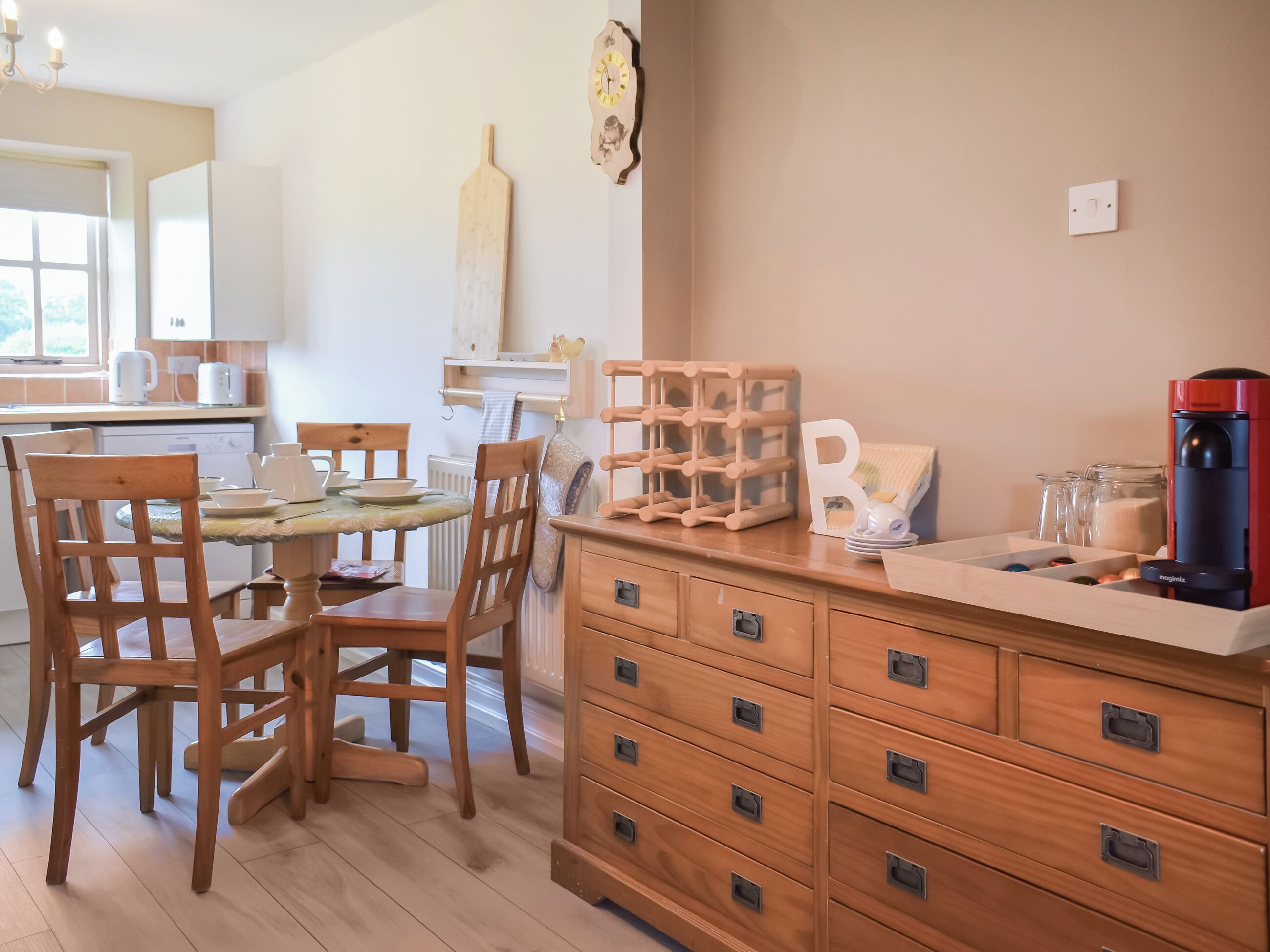 A kitchen with wooden cabinets, a dining table with chairs, a coffee maker, and a clock on the wall.