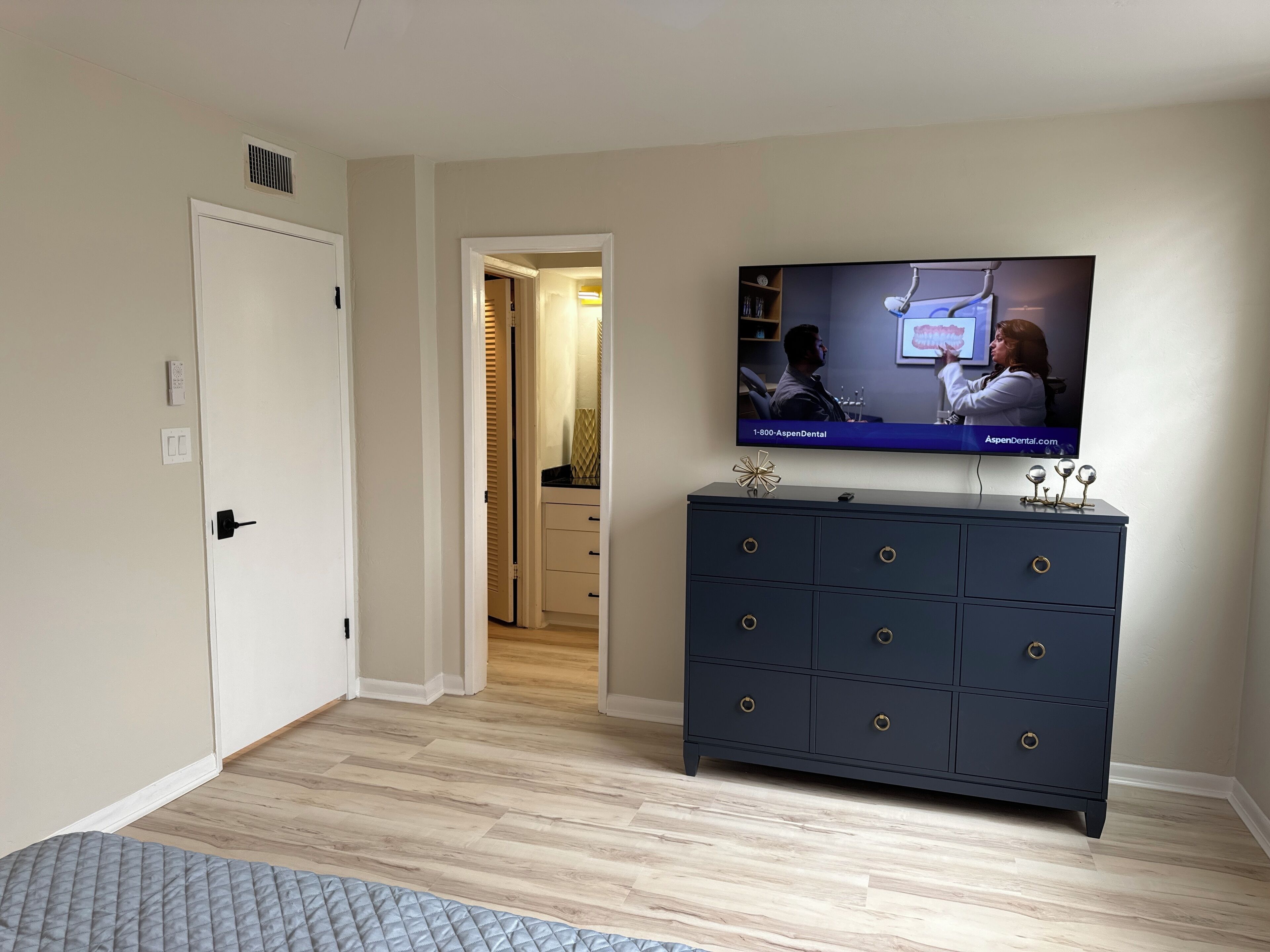 Una habitación con una televisión montada en la pared, un aparador azul oscuro con cajones, suelo de madera y una puerta que da a otra habitación.