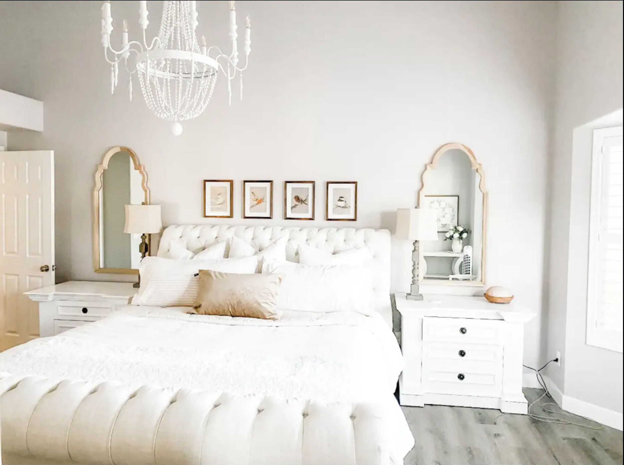A bedroom with a tufted white bed, two bedside tables, a chandelier, and framed pictures on the wall.