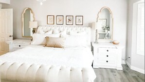A bedroom with a tufted white bed, two bedside tables, a chandelier, and framed pictures on the wall.