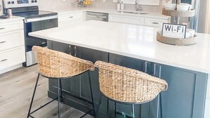 A modern kitchen with white cabinets, a large island with two wicker barstools, a stainless steel refrigerator, and a sign indicating Wi-Fi availability.