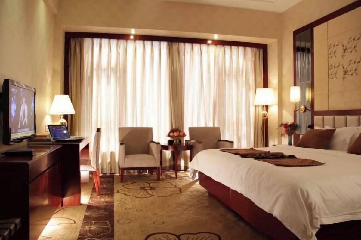 A hotel room with a large bed, two armchairs, a desk with a laptop, a TV, and a large window with curtains.