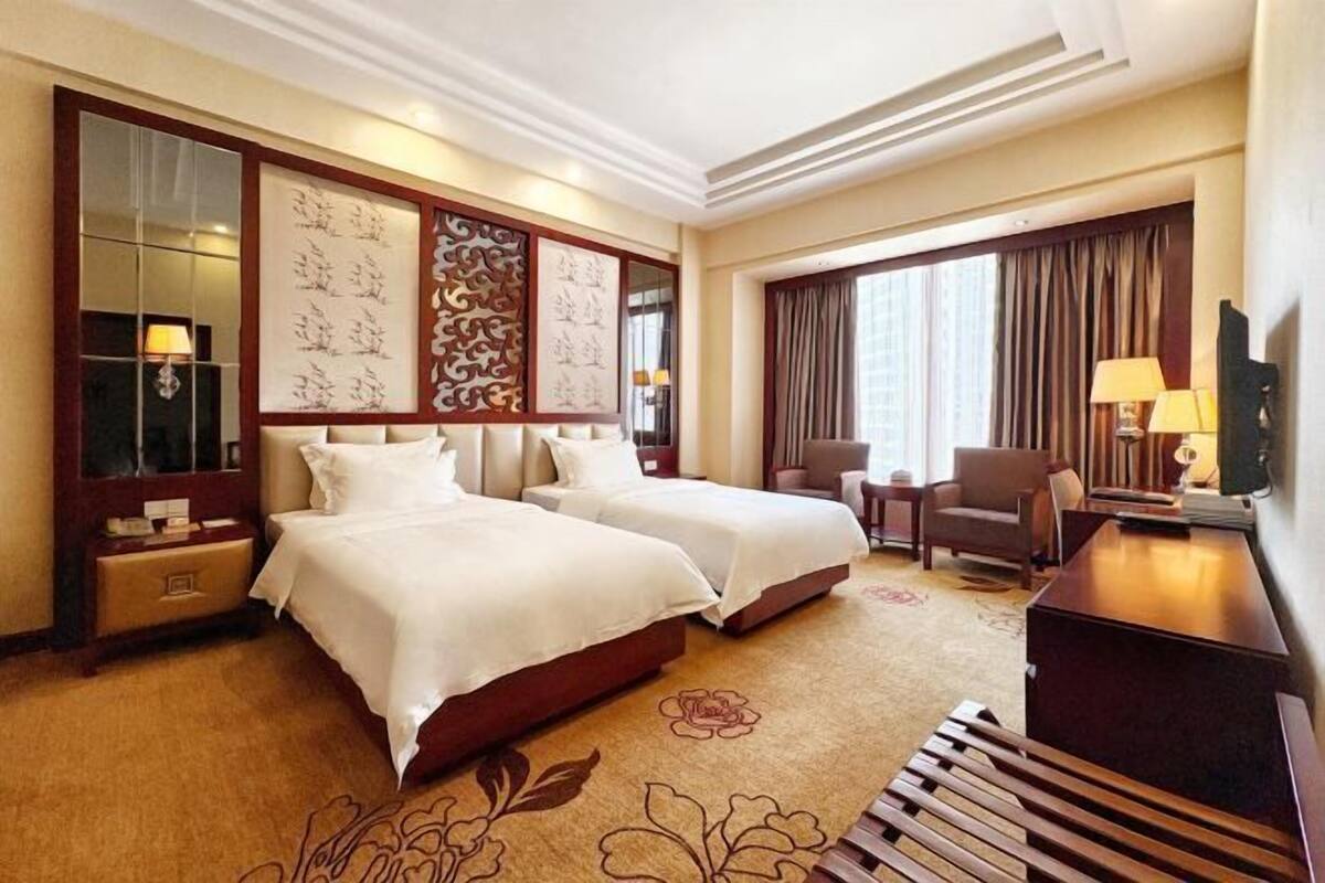 A hotel room with two beds, a desk, a chair, a TV, and a large window with curtains.