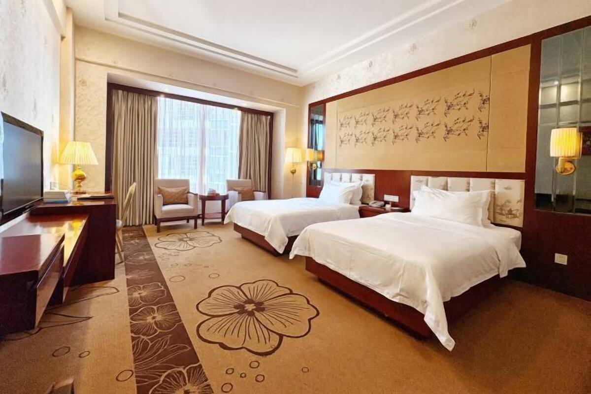A hotel room with two beds, a desk with a TV, a chair, and a large window with curtains.