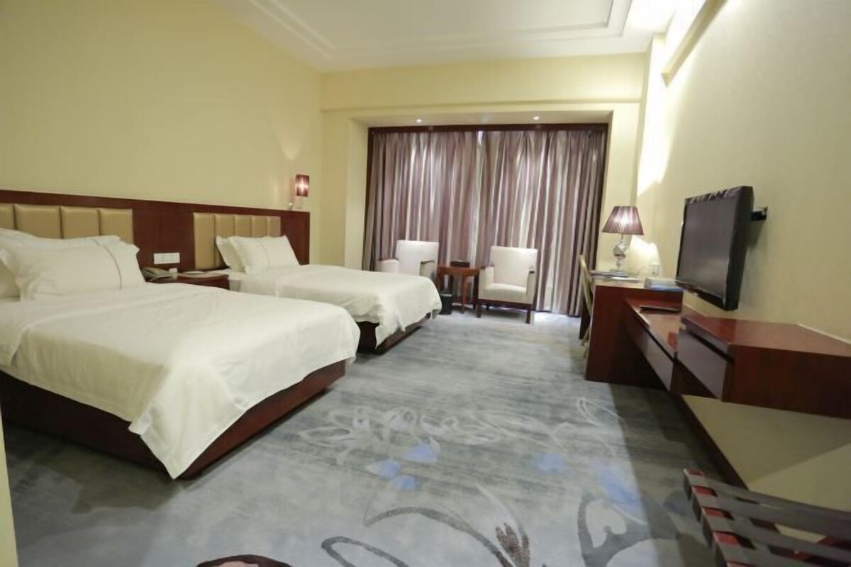 A hotel room with two beds, a desk, a chair, a TV, and a large window with curtains.