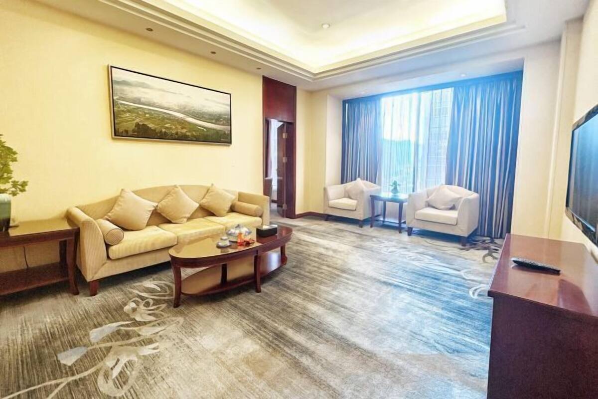 A hotel room with a sofa, armchairs, a coffee table, a TV, and a large window with curtains.