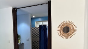 Une chambre d’hôtel avec un sol à motifs, un lit doté d’une tête de lit décorative, un miroir fixé au mur et une vue sur la salle de bain, où l’on aperçoit une cabine de douche à carreaux.