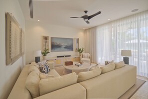 A living room with a large beige sectional sofa, a round coffee table, and a ceiling fan. There are two side tables with lamps, a painting on the wall, and a window with sheer curtains. - Modern Lake Villa With English Speaking Staff in Golf and Beach Resort (Punta Cana)
