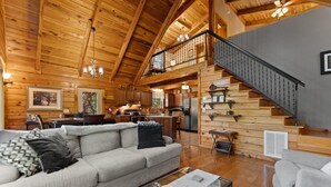 Un salón espacioso con techo alto y vigas de madera, una escalera y una cocina al fondo.