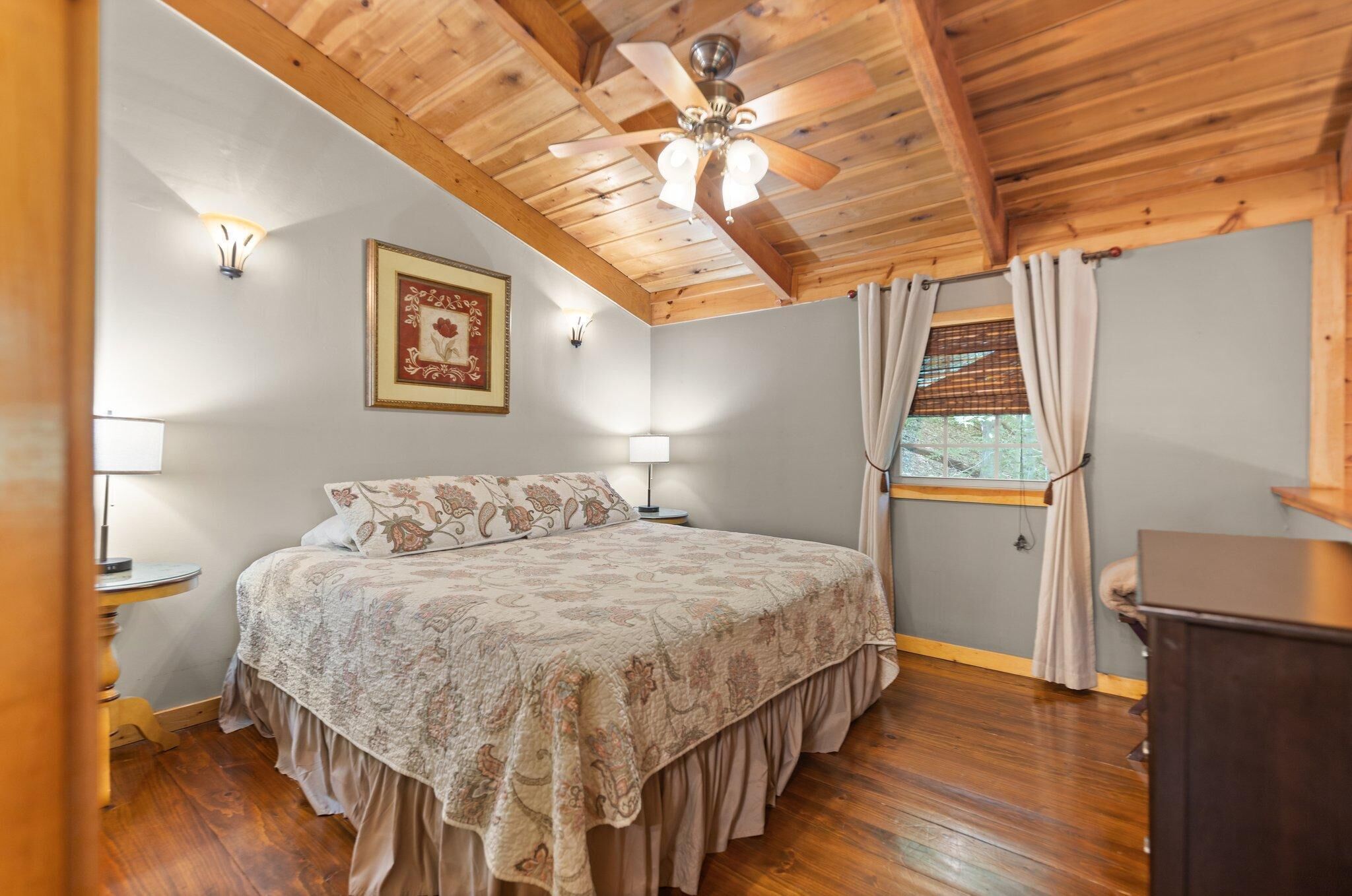 A bedroom with a wooden ceiling, a bed with floral bedding, two bedside lamps, a framed picture on the wall, and a window with curtains.