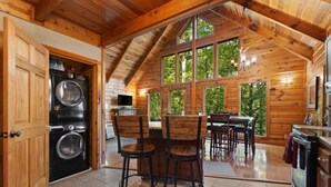 Interior de una cabaña de madera con cocina, comedor y ventanas amplias.