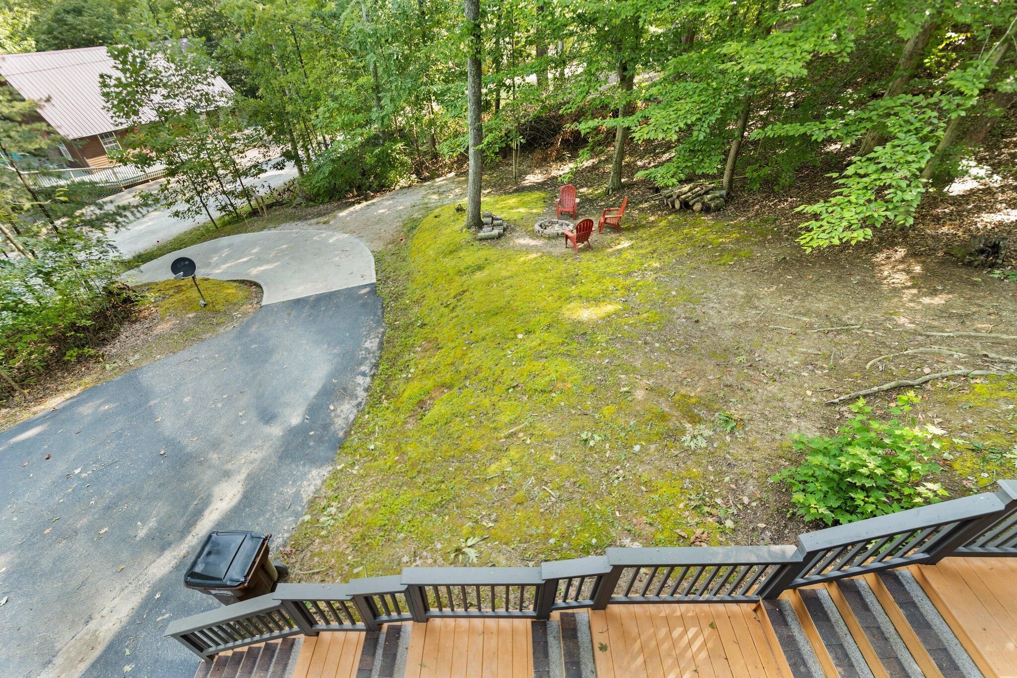 Una terraza de madera con baranda metálica, un sendero pavimentado que lleva a una casa y un espacio con fogata que incluye dos sillas rojas.