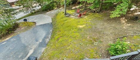 Una terraza de madera con baranda metálica, un sendero pavimentado que lleva a una casa y un espacio con fogata que incluye dos sillas rojas.