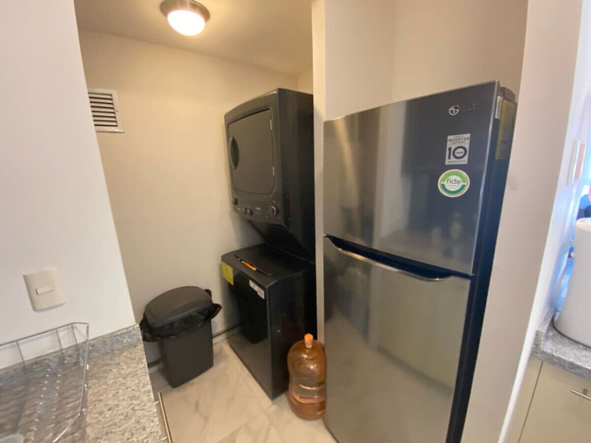 A compact kitchen with a stainless steel refrigerator, a small trash bin, and a water dispenser.