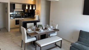 Una cocina moderna con electrodomésticos integrados, un comedor con mesa de madera y sillas blancas, y un sofá gris en la sala.