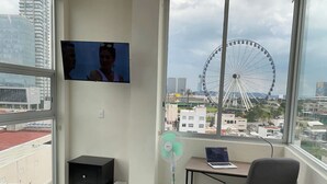 A hotel room with a bed, a desk with a laptop, a chair, a TV mounted on the wall, and a large window with a view of a Ferris wheel and buildings. - NEW Penthouse for 8 in La Estrella de Puebla (Heroica Puebla de Zaragoza)