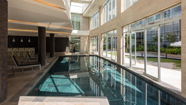 Una piscina interior moderna con paredes de vidrio transparente, ventanas amplias y un interior iluminado por el sol.