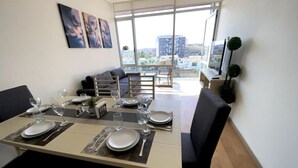 Un comedor con una mesa preparada para cuatro personas, un sofá y vistas a la ciudad a través de amplios ventanales.
