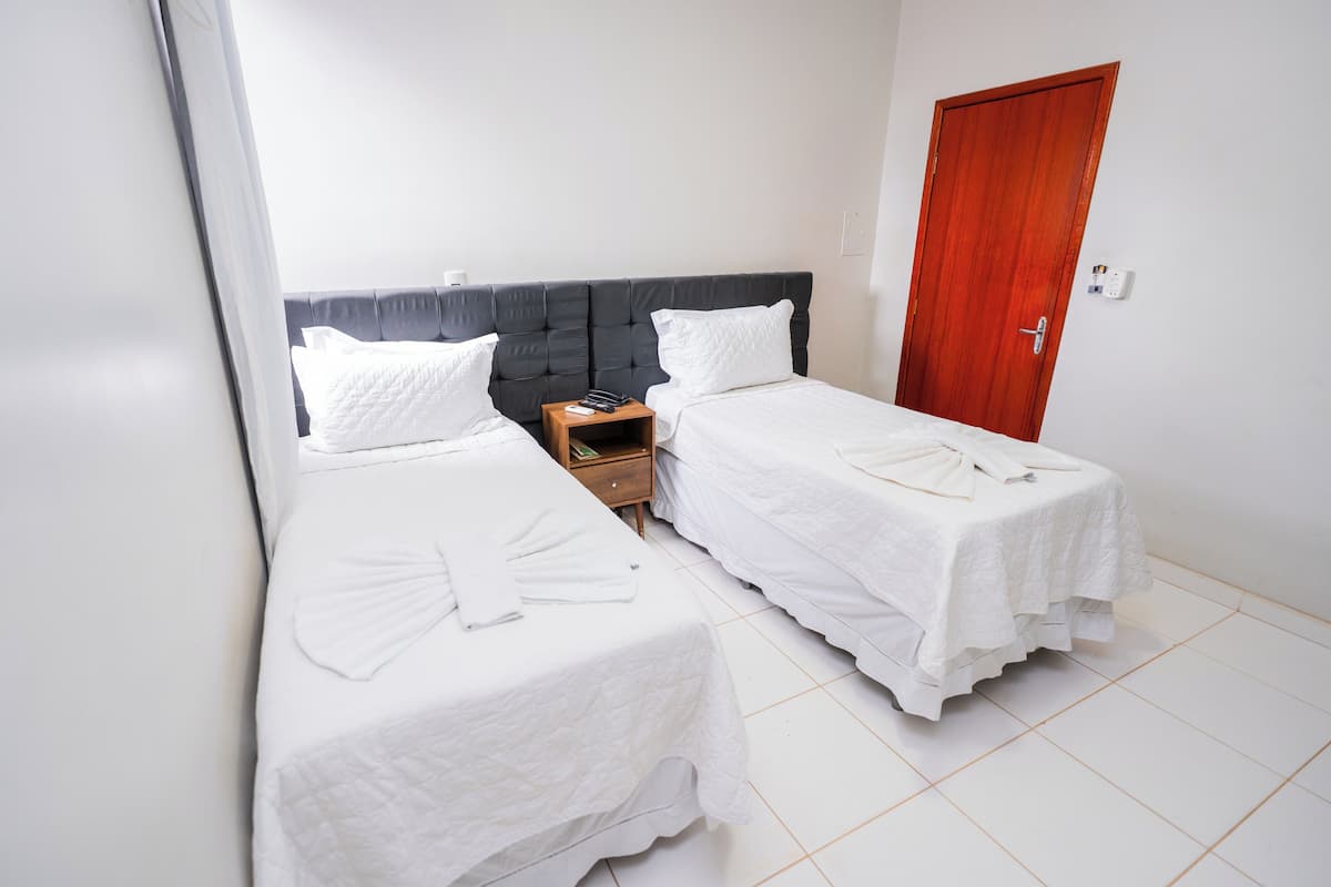 A hotel room with two single beds, a wooden nightstand, a wooden door, and white tiled flooring.
