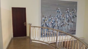 A hallway with a dark wooden door, a metal staircase railing, and a large painting of mounted soldiers on horseback. - Sultanate Suites GRA Kano (Kano)