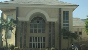 A building with a large arched window, a tiled roof, and a paved entrance area with potted plants. - Sultanate Suites GRA Kano (Kano)