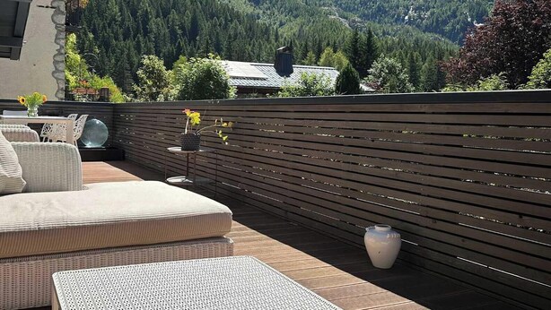 Una terraza de madera con mobiliario exterior, una mesa de vidrio y una planta en maceta.