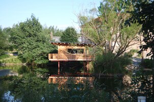 A wooden houseboat with a deck and a small cabin, surrounded by trees and a calm body of water.