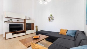 Un salon moderne doté d’un téléviseur à écran plat, d’une table basse en bois, d’un canapé gris foncé agrémenté de coussins et d’un luminaire fixé au mur.