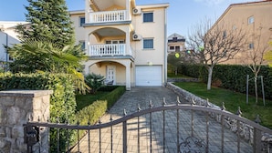 Un immeuble résidentiel de deux étages doté d’un balcon central, d’un garage et d’une allée pavée menant à une entrée fermée par un portail.