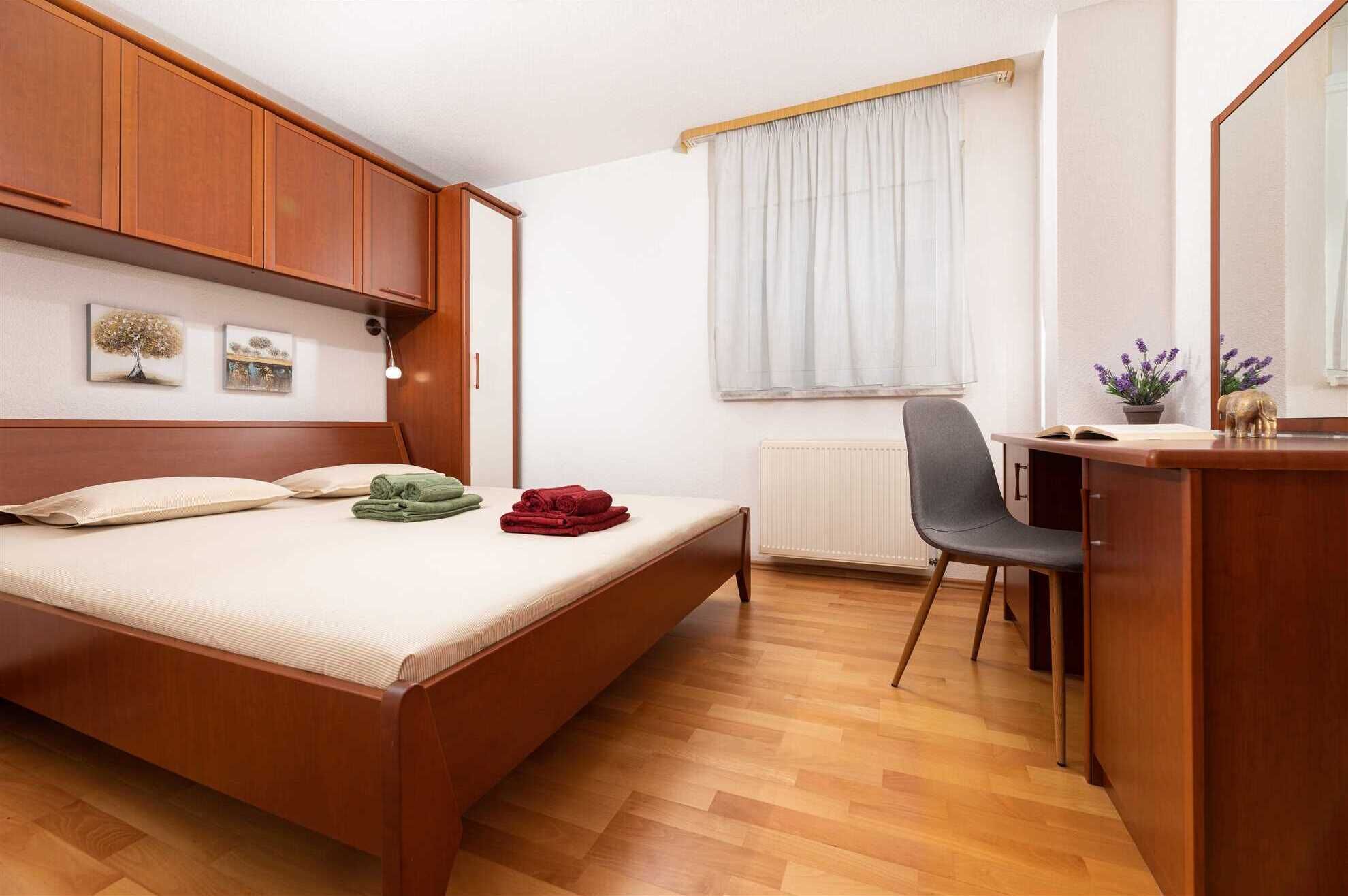 A bedroom with a wooden bed, a desk with a chair, a radiator, and a window with curtains.
