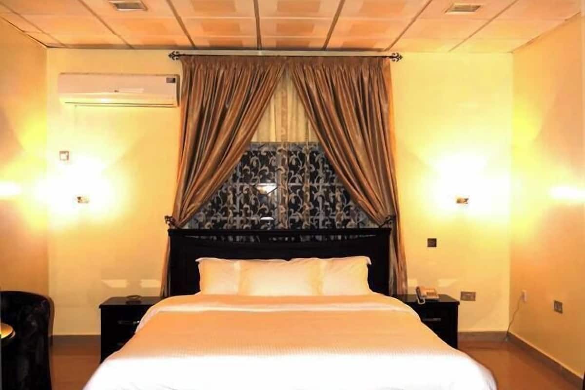 A hotel room with a large bed, a headboard with a decorative pattern, two bedside lamps, and a window with curtains.