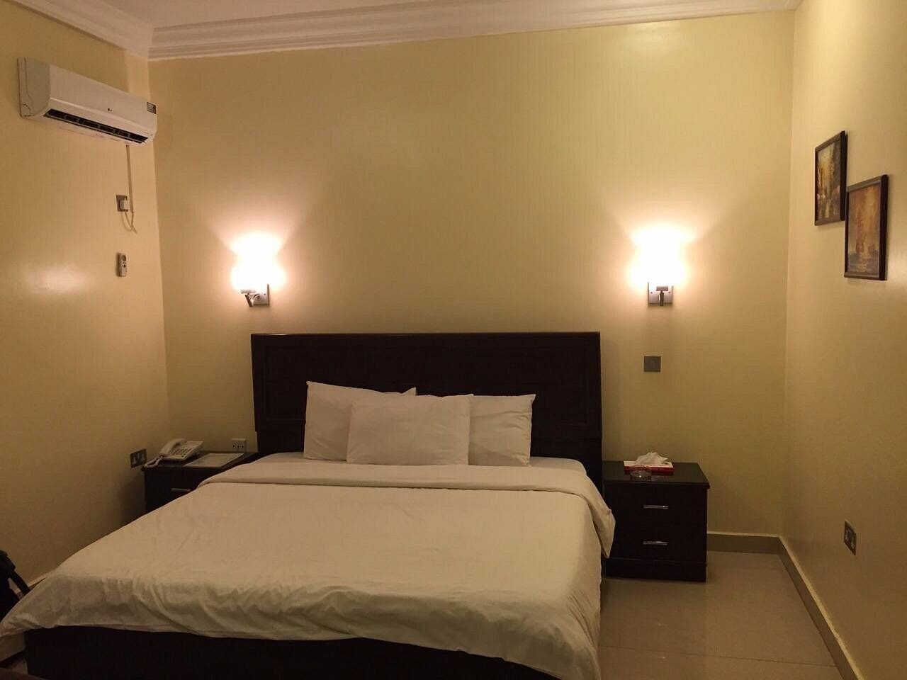 A hotel room with a bed, bedside tables, an air conditioning unit, and wall-mounted lights.