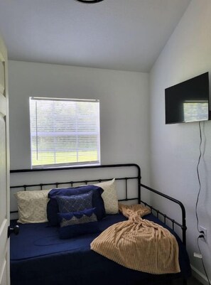 Une petite chambre avec un lit simple, une fenêtre, un téléviseur à écran plat fixé au mur et une prise de courant sur le côté.