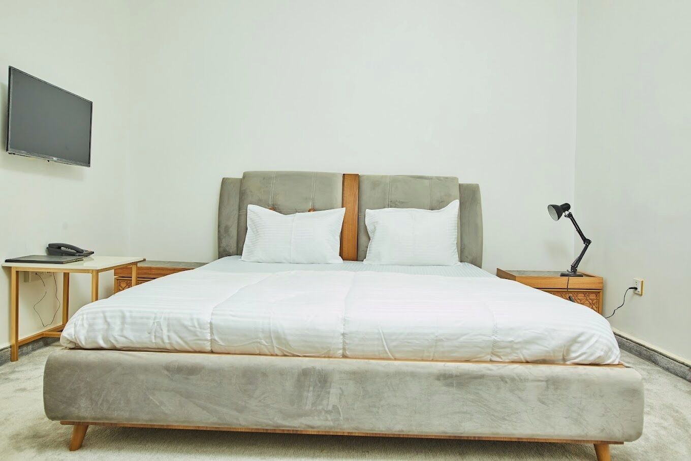 Een keurig opgemaakt bed met een grijs beklede hoofdbord, een nachtkastje met een lamp, en een flatscreen televisie aan de muur bevestigd.