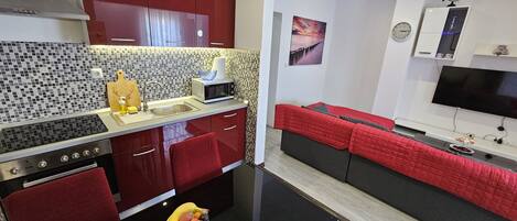 A compact kitchen with a central island, a dining area with a red sofa, and a flat-screen TV mounted on the wall.