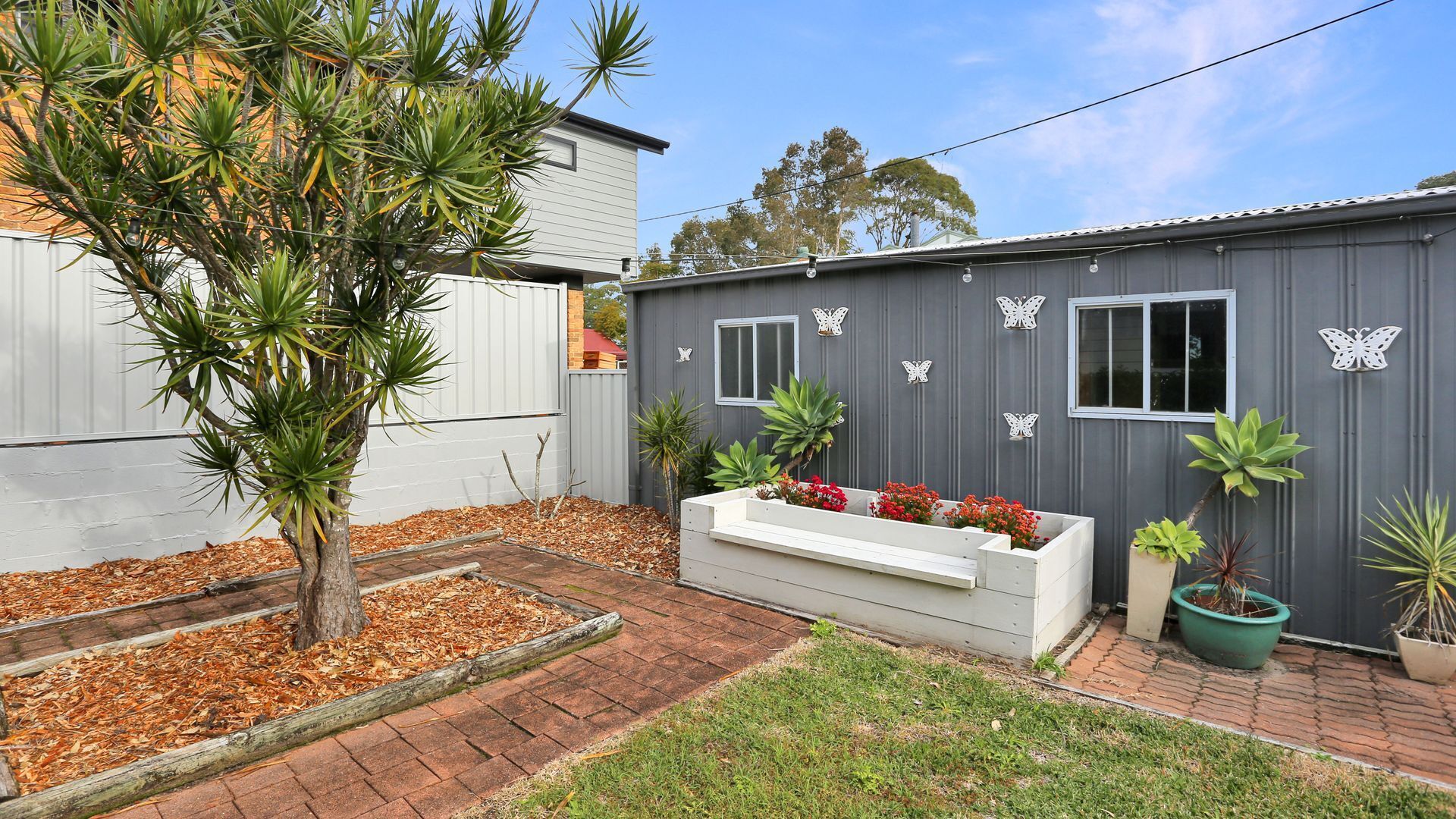 A residential property with a grey shed, a white bench with red flowers, and various potted plants.