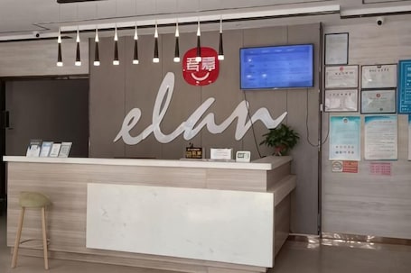 A reception desk with a sign that reads 'elan,' a television mounted on the wall, and a bulletin board with various notices.. Junjia Hotel (Ji'an Pedestrian Street)