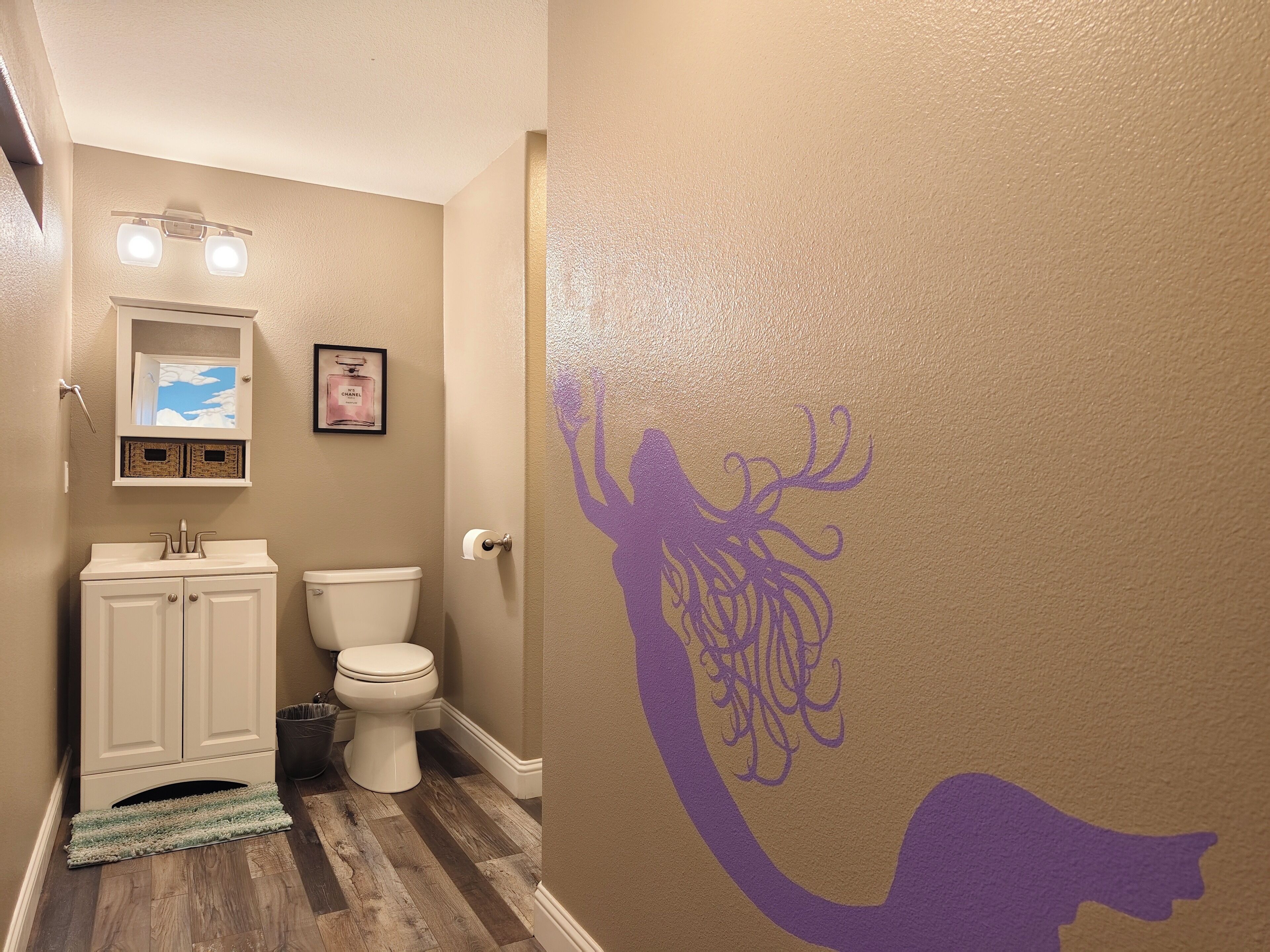 Une salle de bain avec un plancher en bois, des meubles blancs, des toilettes et un autocollant mural représentant une sirène.