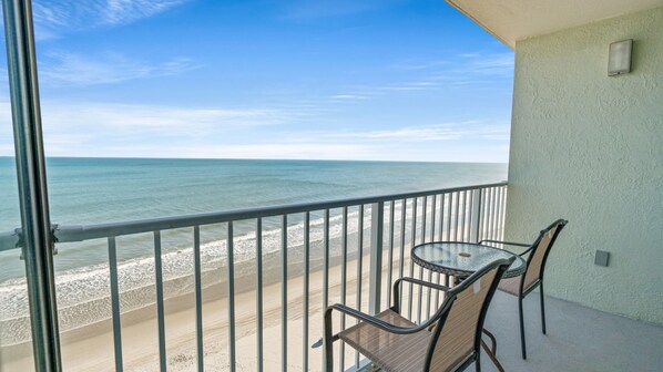 Ein Balkon mit Glastür, einem kleinen Tisch und zwei Stühlen mit Blick auf einen Strand.