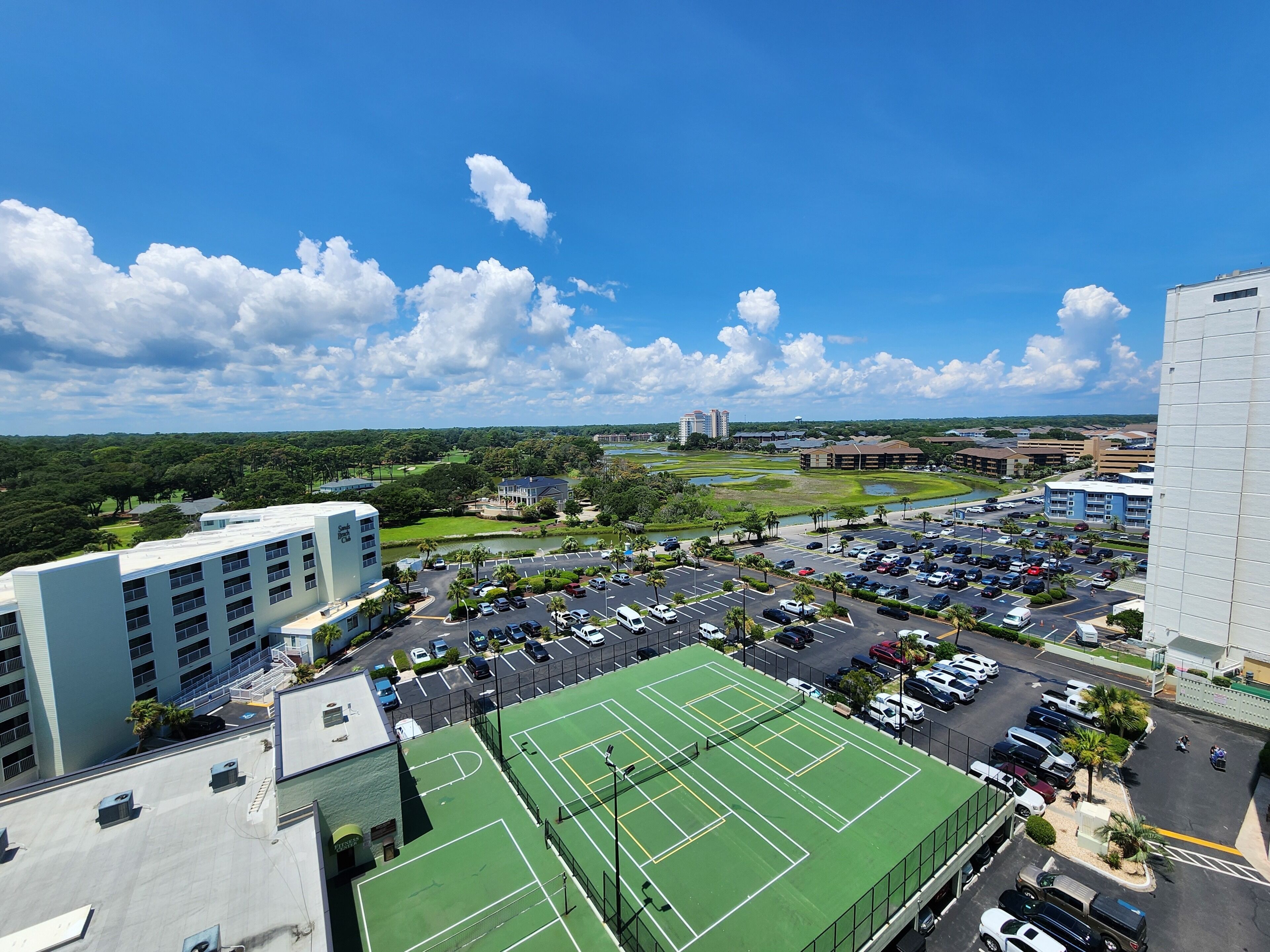 A multi-story hotel with a rooftop tennis court, parking lot, and a view of a golf course and cityscape.