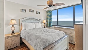 A bedroom with a bed, two bedside tables, a ceiling fan, a window with a view of the ocean, and a nightstand with a lamp.