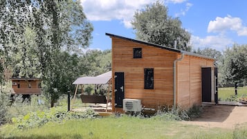 A wooden cabin with a single door and a small window, surrounded by greenery and trees.