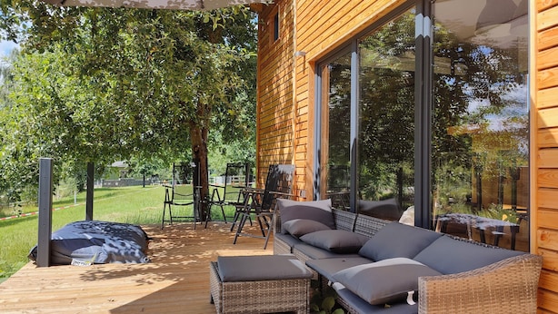 Un terrazzo in legno con divano e poltrone, ampie finestre e vista su un paesaggio verde.
