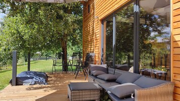 A wooden deck with a sofa and chairs, large windows, and a view of a green landscape.