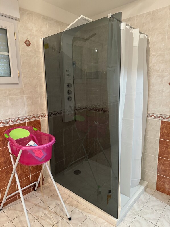 A glass-enclosed shower stall with a showerhead and control knobs, a pink plastic basin on a white stand, and a window with a frosted glass panel.