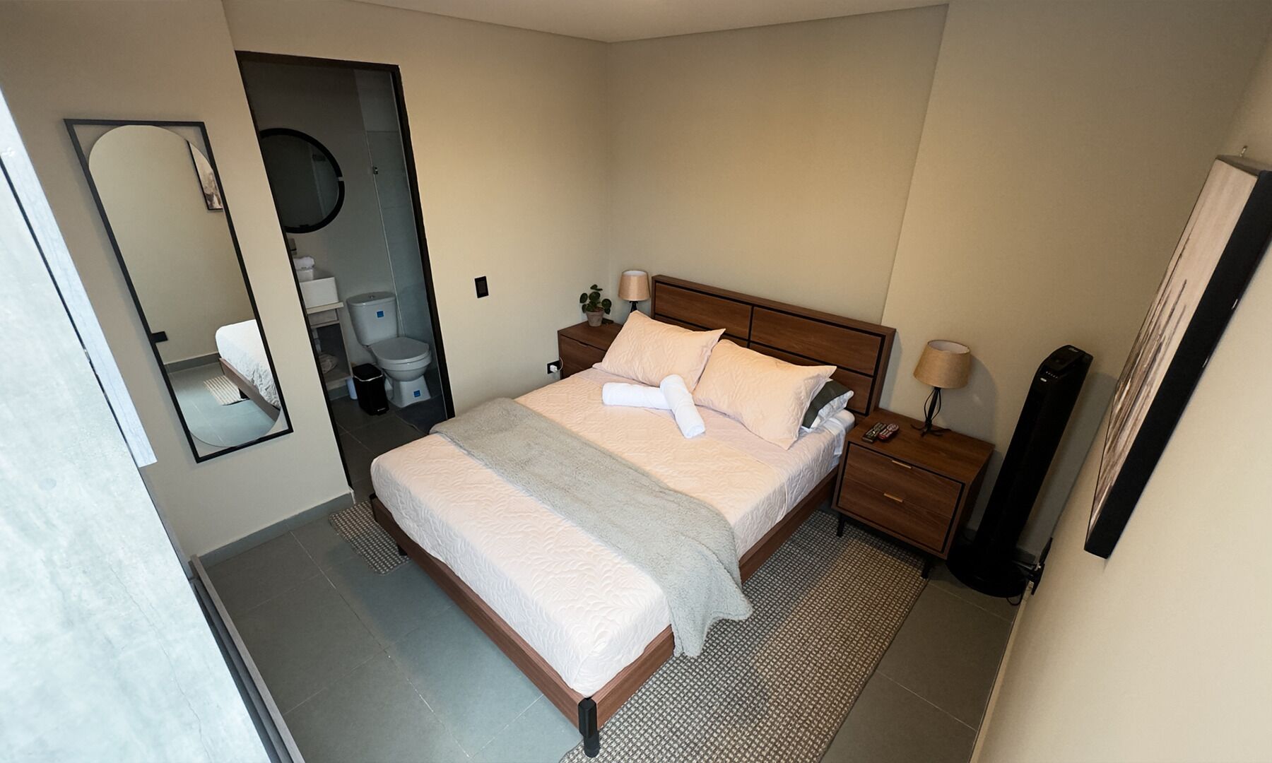Ein ordentlich eingerichtetes Hotelzimmer mit einem Bett, Nachttischen, einem Spiegel und einem durch eine offene Tür einsehbarer Badezimmer.