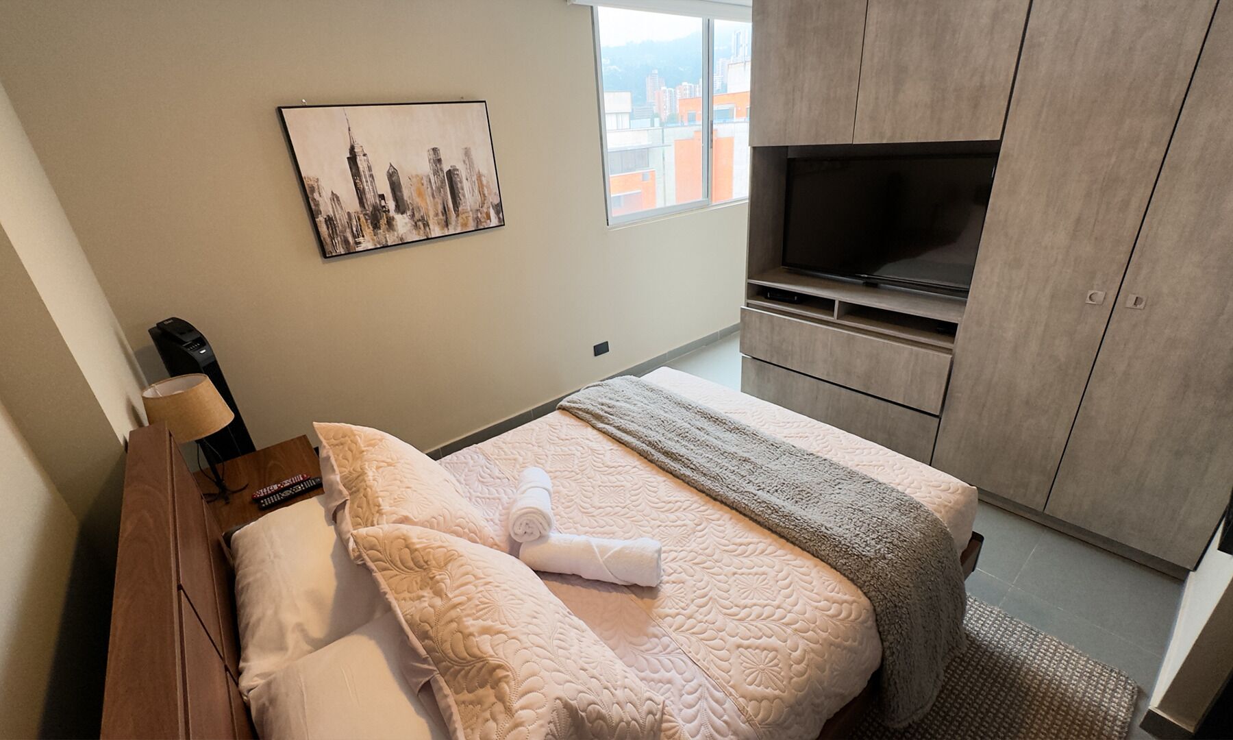 A hotel room with a bed, a TV, a painting, and a window with a view of buildings.